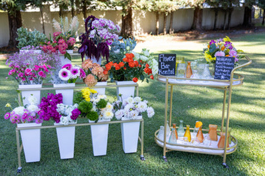 Outdoor setting with floral arrangements in white pots and a bar cart with drinks on grass.