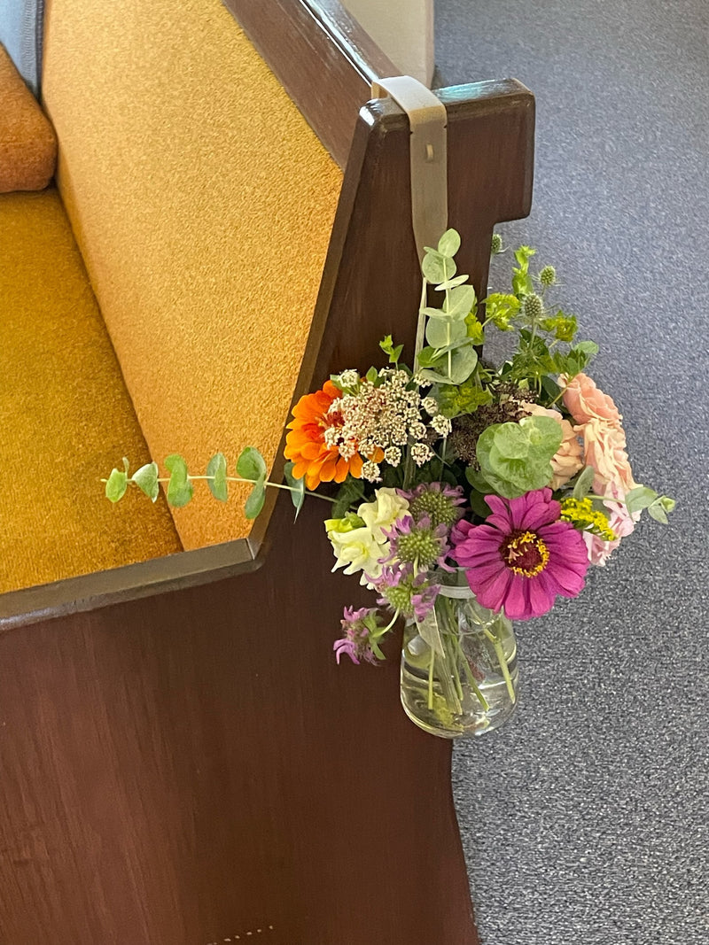 Bouquet of flowers in a clear vase attached to a wooden church pew.