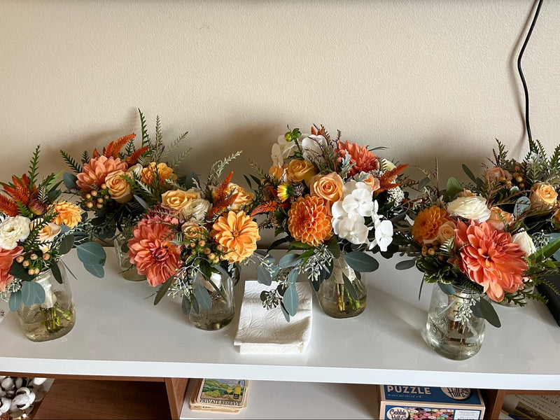 Row of small floral arrangements in glass vases on a table with a TV in the background.