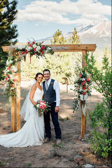 Wedding couple standing under a wooden arch decorated with flowers in a natural setting.