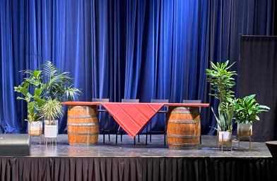 Decorative setup with barrels, a red tablecloth, and plants against a blue curtain.