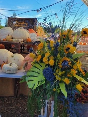 Floral arrangement with sunflowers and blue flowers in an outdoor setting with pumpkins and a blue sky.