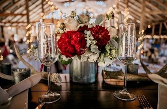 Decorative table setting with red flowers, wine glasses, and candles in a warm indoor setting.