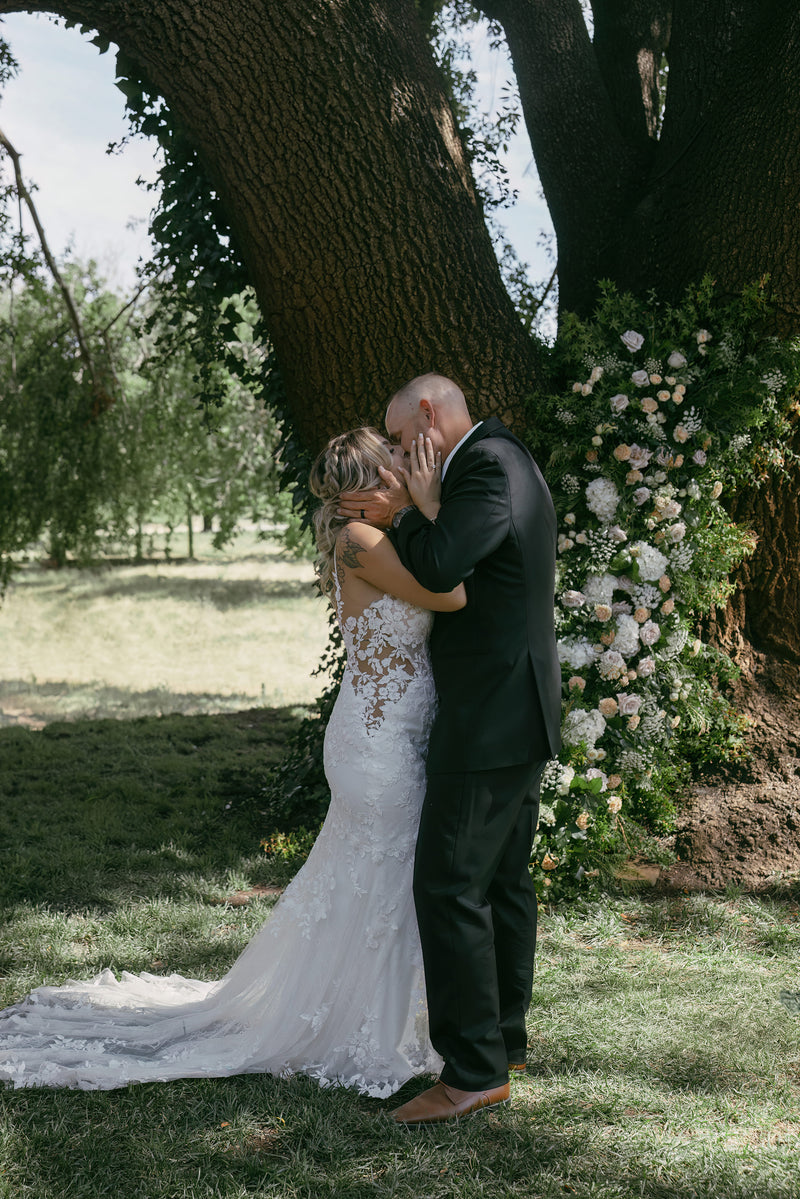 Couple embracing under a large tree with floral decorations on a sunny day.