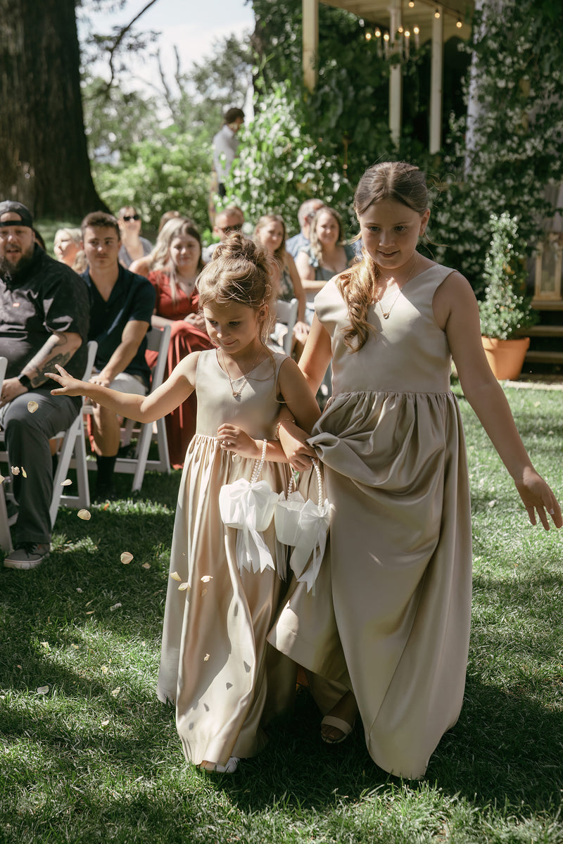 Two young girls in formal dresses walking outdoors with a crowd in the background.