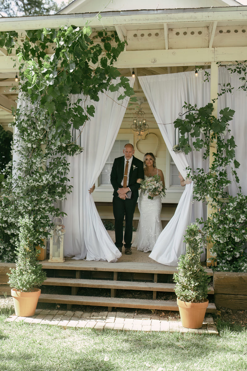 Wedding couple standing under a decorated archway with greenery and white curtains on a wooden porch.
