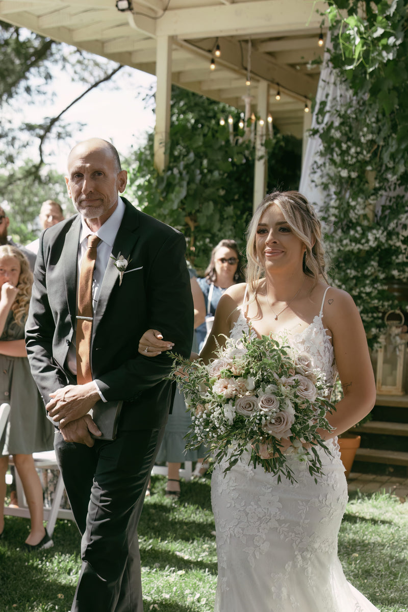 Bride walking down the aisle with a man, surrounded by greenery and people.